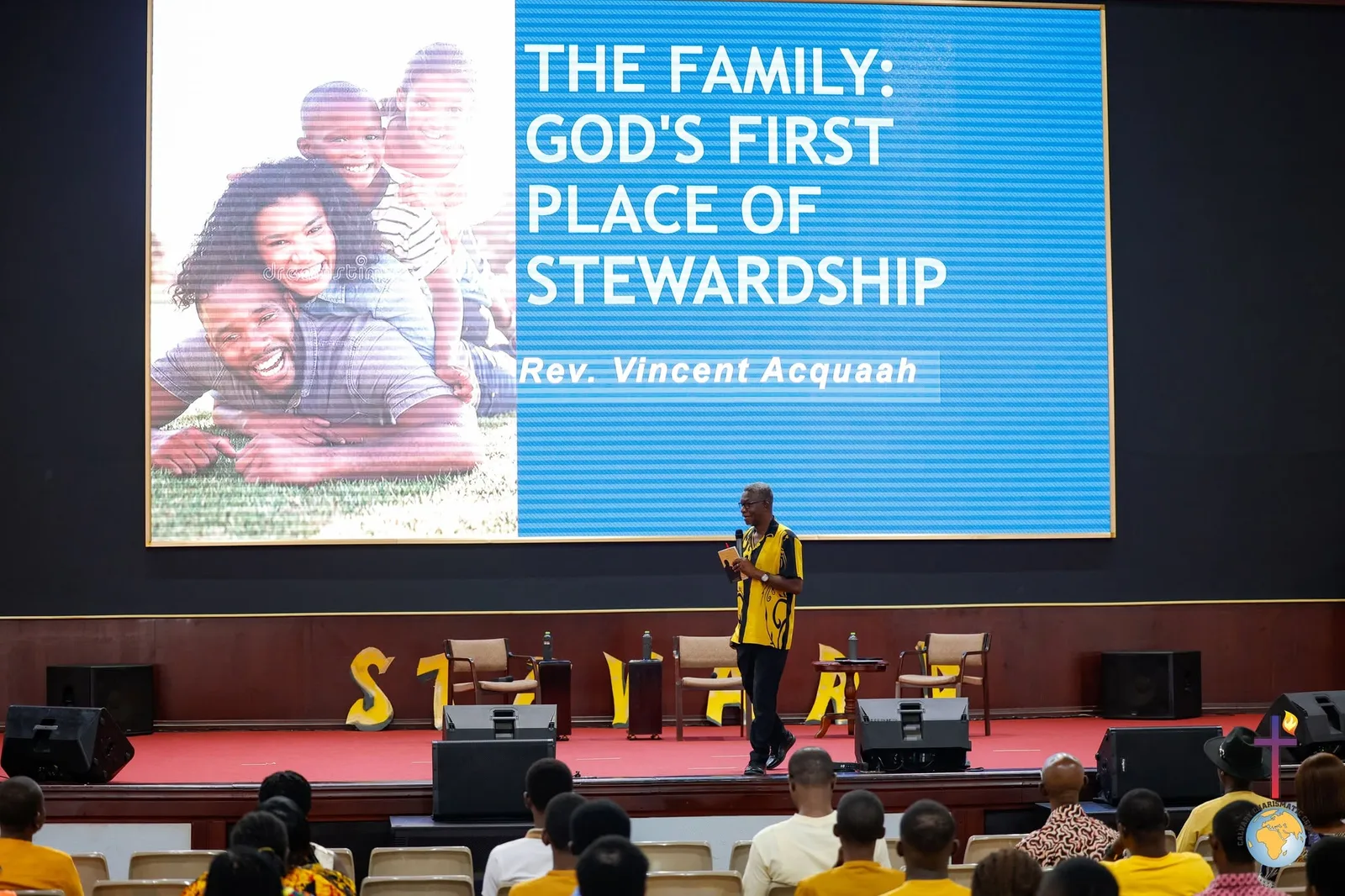 Rev. Vincent Acquaah on stage in front of a slide reading The Family: God's First Place of Stewardship