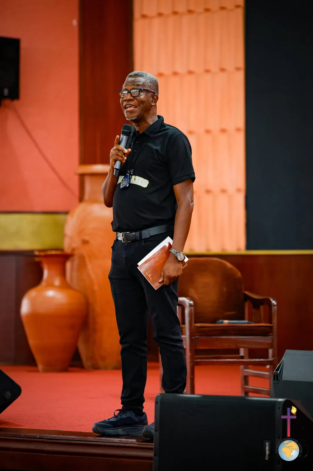 Rev. Vincent Acquaah speaking on stage in front of a clay pot display